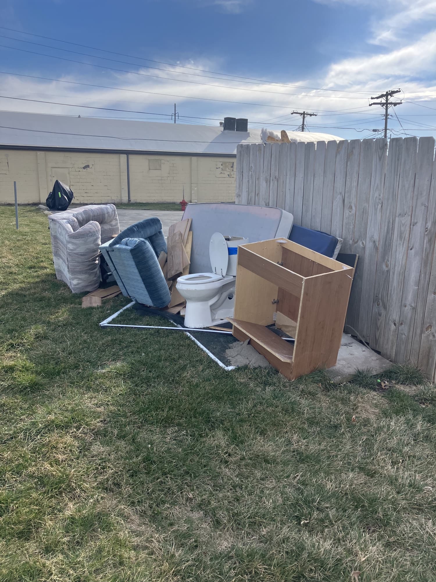 Outdoor eviction set-out pile with furniture, cabinets, and bathroom fixtures staged by a fence.